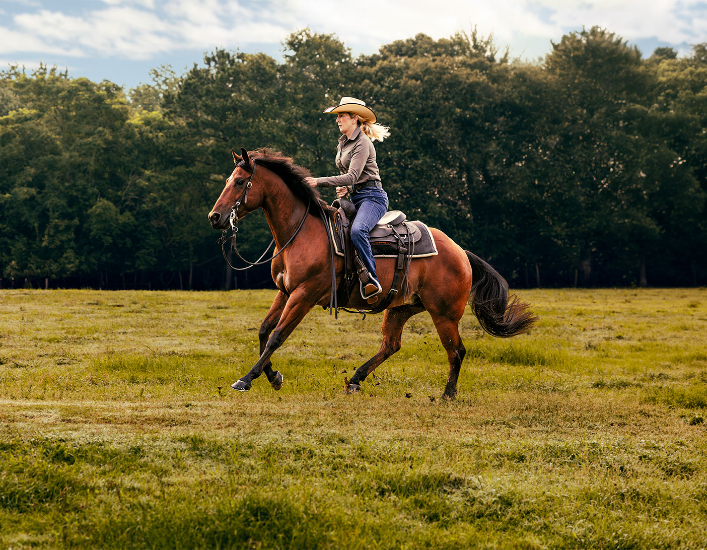 Cowgirl riding a brown horse at a gallop across a grassy field with trees in the background.
